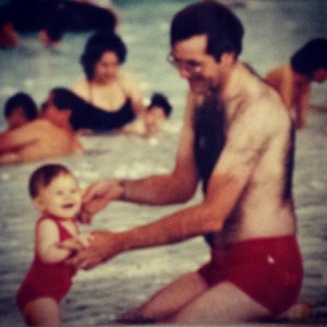 A white-skinned, brunette man with red swim trunks and trucker's tan holds up a tiny human in a red bathing suit. They're in shallow water of a pool. Other swimmers can be seen in the background.