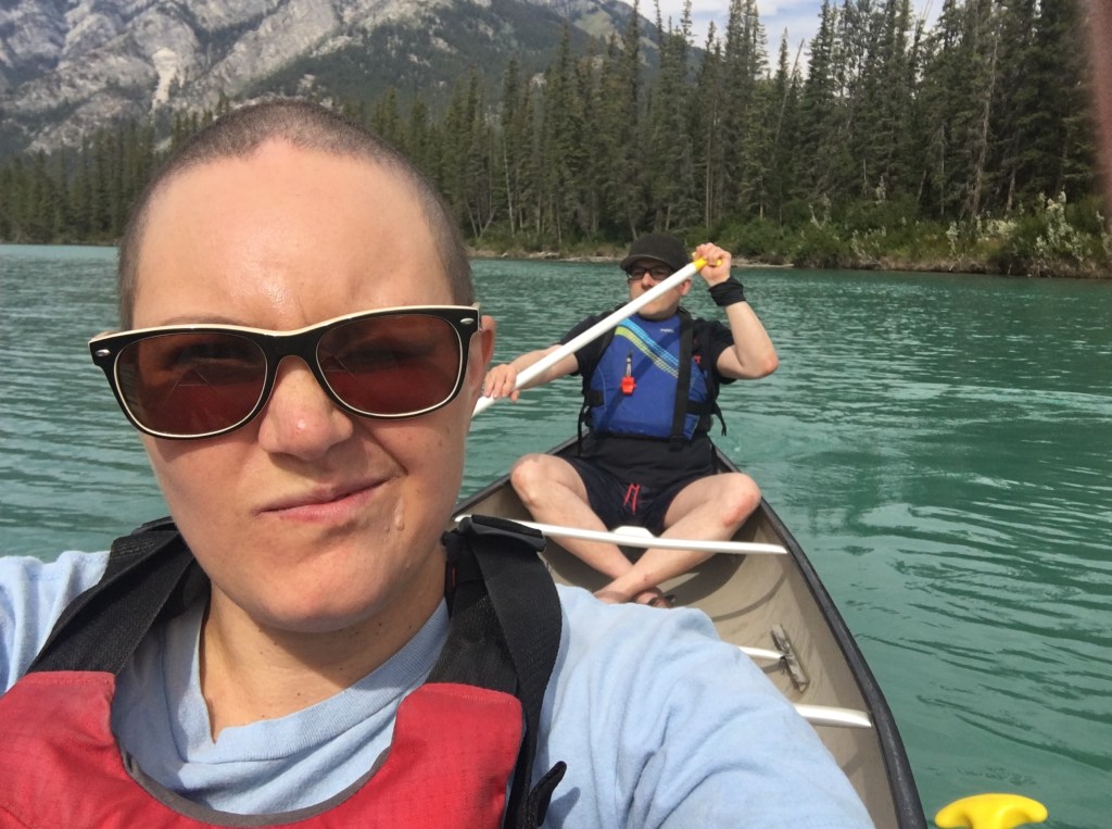 A short-haired (almost bald), peach-skinned person in sunglasses smirks into the camera while a blue-life jacketed man with a black cap paddles the canoe. They float above turquoise water with a rocky mountain and spruce forest in the background.